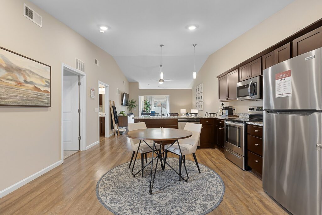 Open-concept kitchen and dining area with stainless steel appliances, dark wood cabinets, round table on a rug, and light wood flooring. Living area is visible in the background.