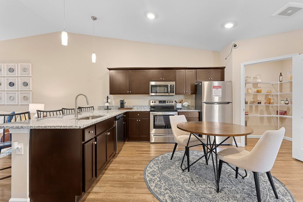 Modern kitchen with dark wood cabinets, stainless steel appliances, granite countertops, a round dining table with two chairs, and a pantry with shelves visible through an open door.