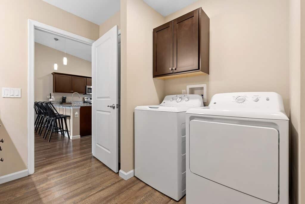 A laundry room with a white washer and dryer, brown cabinets above, and a view into a kitchen with an island and barstools.