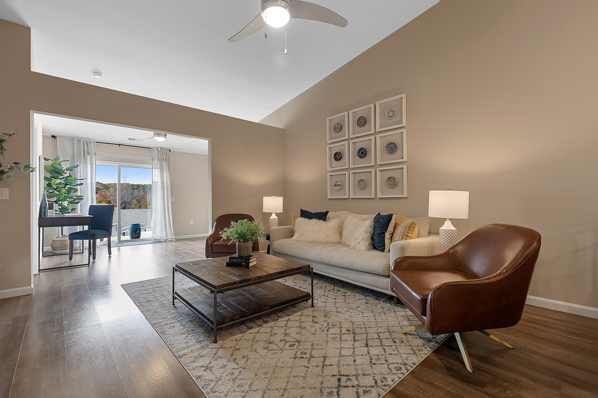 Modern living room with a beige sofa, two brown chairs, coffee table, area rug, and wall art. A separate office space with a desk and window view is visible in the background.
