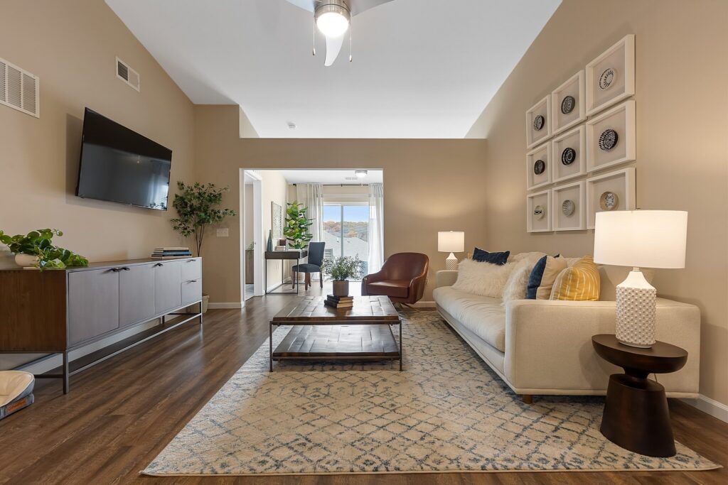 Modern living room with a beige sofa, coffee table, patterned rug, wall-mounted TV, plants, and decorative plates on the wall; a dining area is visible in the background.