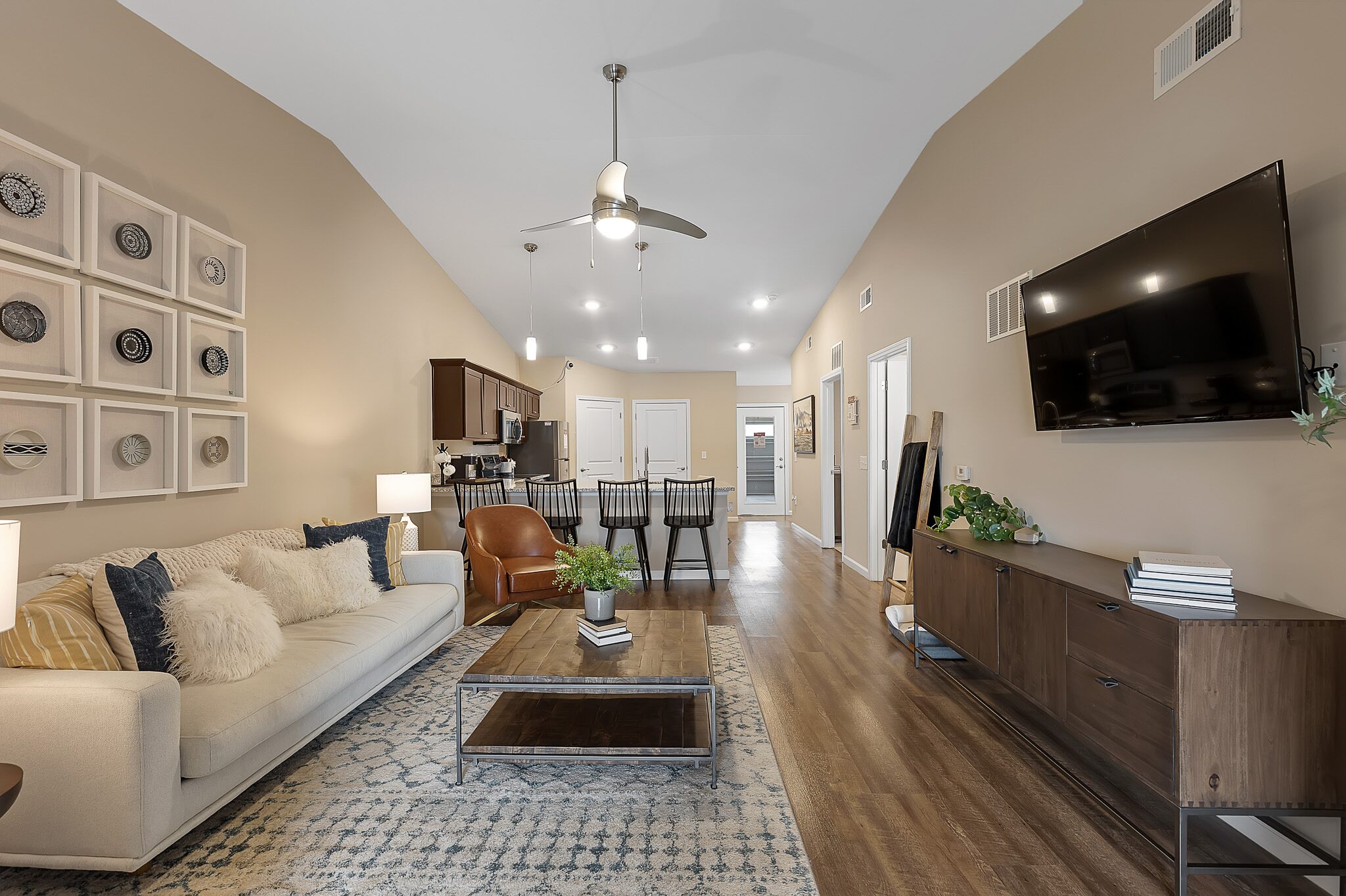 Modern living room with beige walls, a sofa, armchair, coffee table, wall-mounted TV, and dining area with kitchen in the background; wood flooring and contemporary decor.
