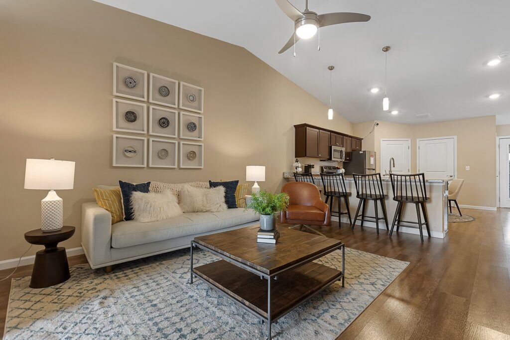 Open-concept living room and kitchen with a beige sofa, patterned rug, coffee table, bar stools, pendant lighting, and neutral decor.