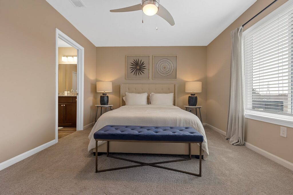A neatly made bed with a tufted headboard sits between two lamps in a beige bedroom; a blue bench is at the foot of the bed, and a window with curtains is on the right.