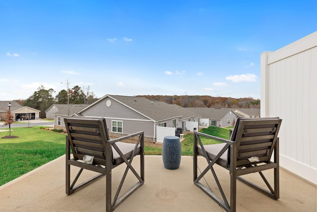 Two wooden chairs and a small table on a patio overlook a row of single-story houses and a grassy yard under a clear blue sky.