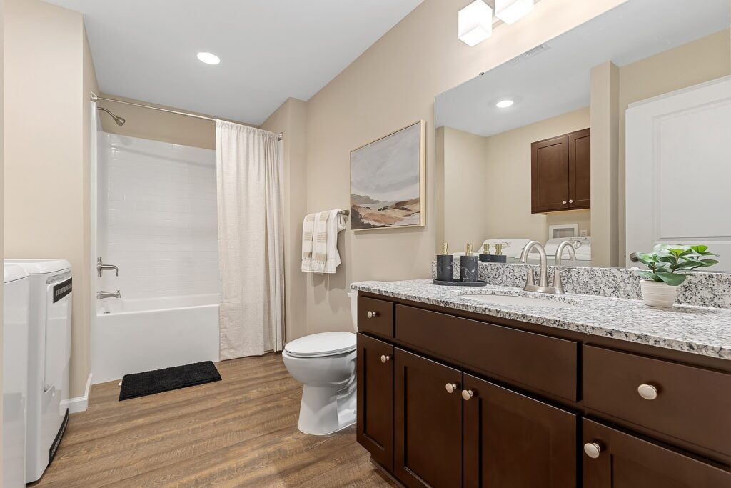Modern bathroom with brown cabinets, granite countertop, toilet, bathtub with shower and curtain, wall art, towel rack, and wood-look flooring.