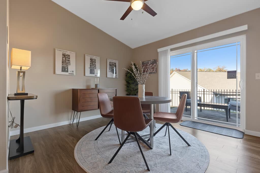 A dining area with a round table and four brown chairs on a circular rug, adjacent to a sliding glass door leading to a balcony with a view of neighboring rooftops.