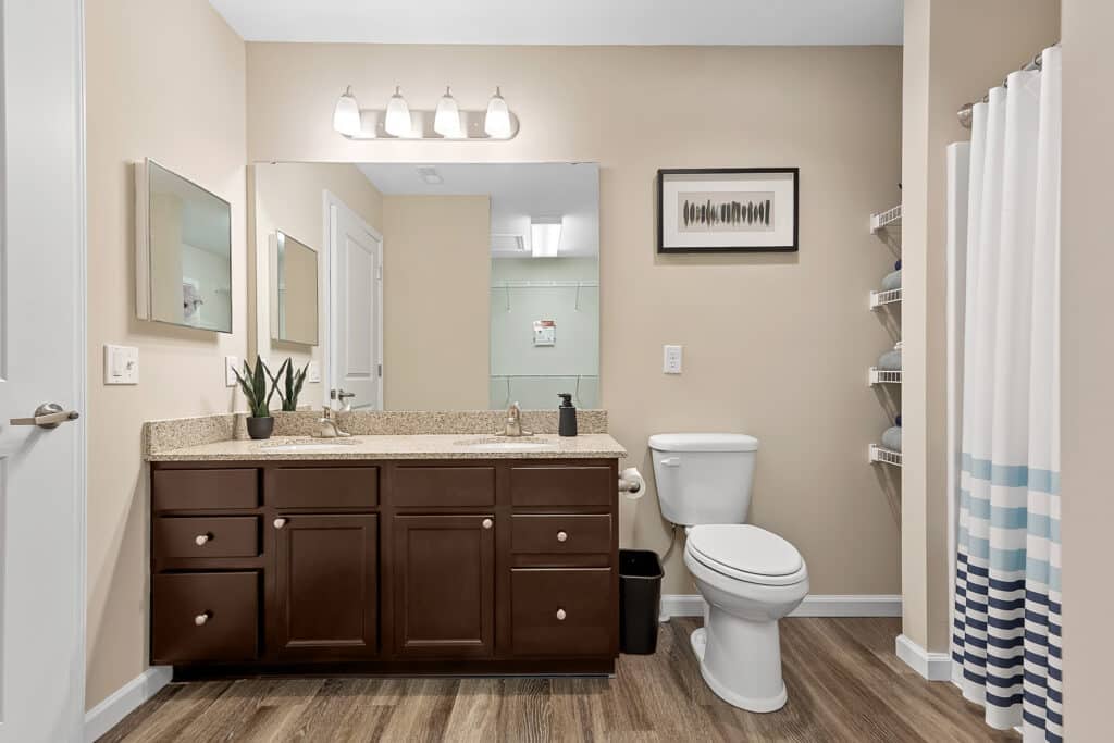Modern bathroom with a brown vanity, granite countertop, large mirror, toilet, shower with striped curtain, wall art, and wooden flooring.