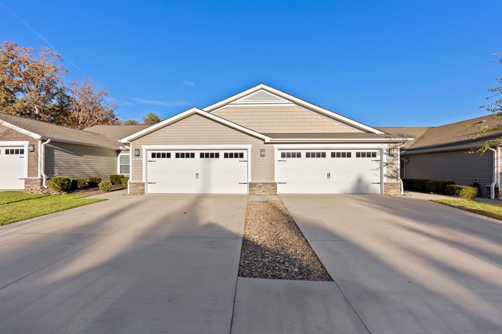 Duplex with two double garages and a shared driveway, surrounded by trimmed grass and shrubs under a clear blue sky.