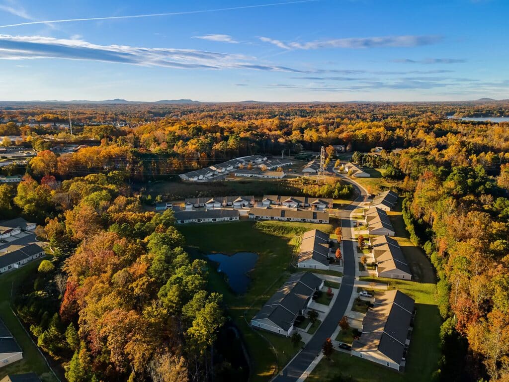 Aerial view of a suburban neighborhood with houses, winding roads, a pond, and surrounding trees with autumn foliage under a clear blue sky.