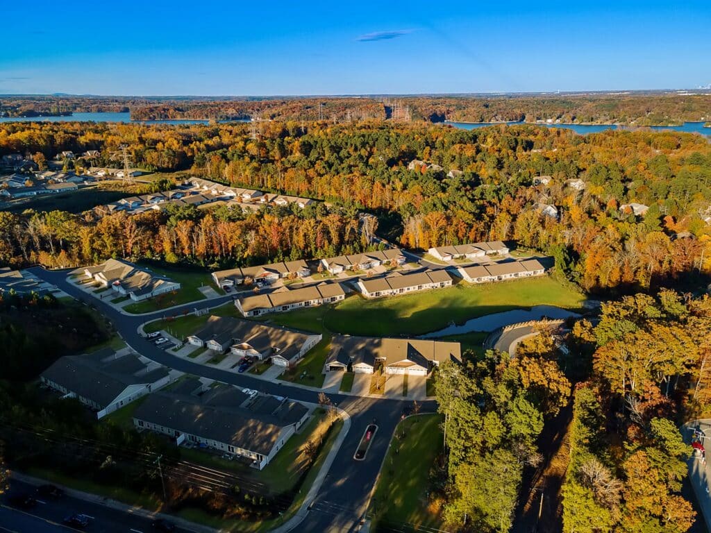 Aerial view of a suburban neighborhood surrounded by trees with autumn foliage, winding roads, and a body of water in the background.