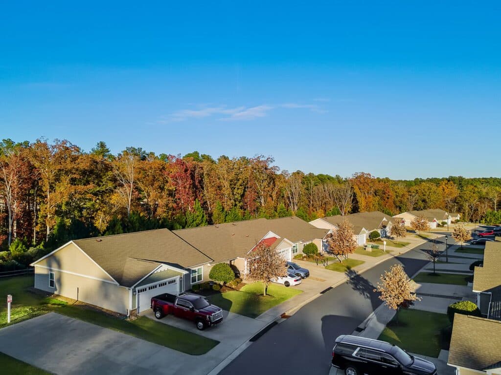 A row of single-story suburban houses with driveways and cars, bordered by a tree line with autumn foliage under a clear blue sky.