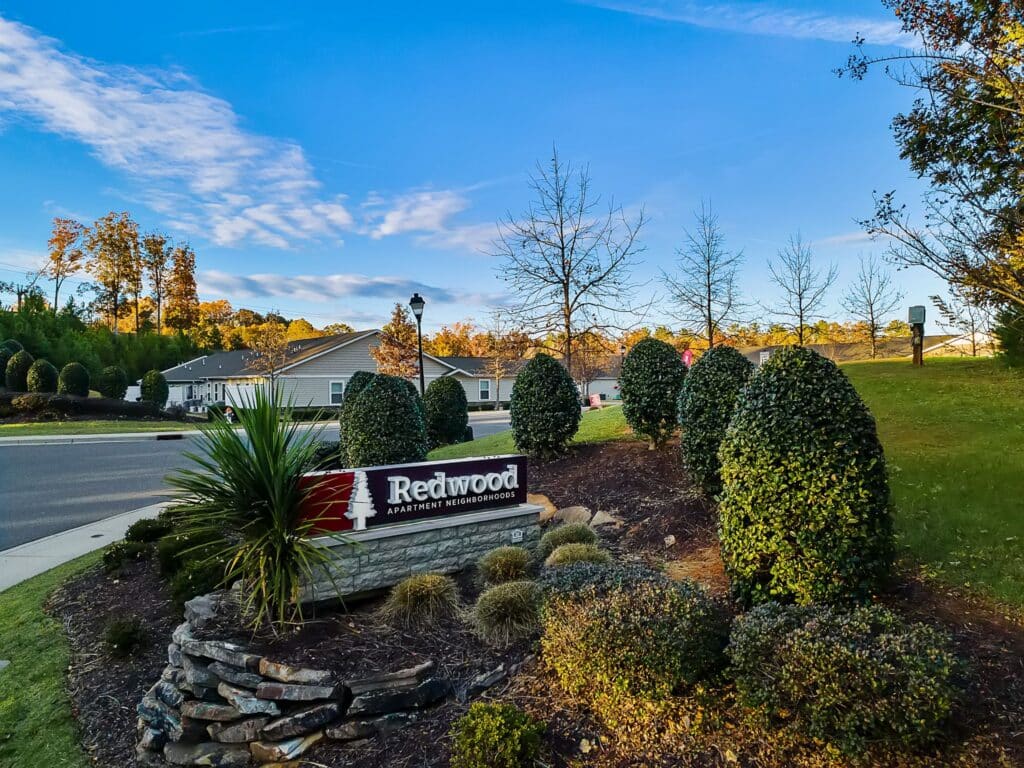 Redwood Apartment Neighborhood sign surrounded by landscaped bushes and plants, with residential houses and a clear sky in the background.