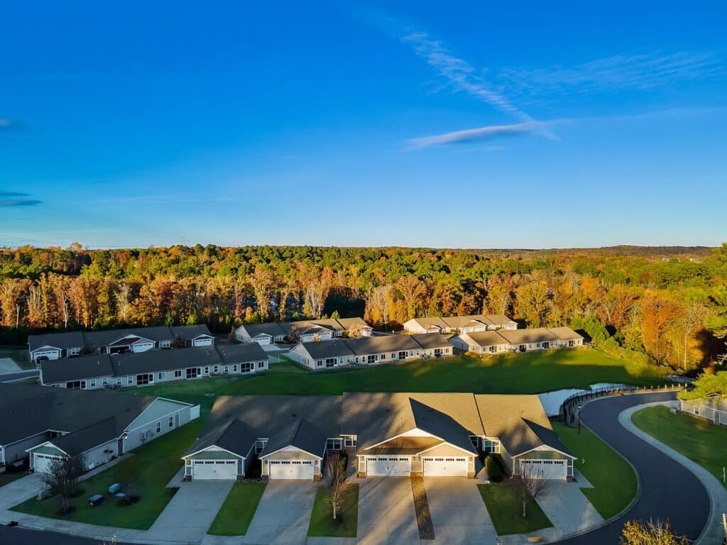 Aerial view of a suburban neighborhood with single-story houses, green lawns, and a tree-lined background under a clear blue sky.