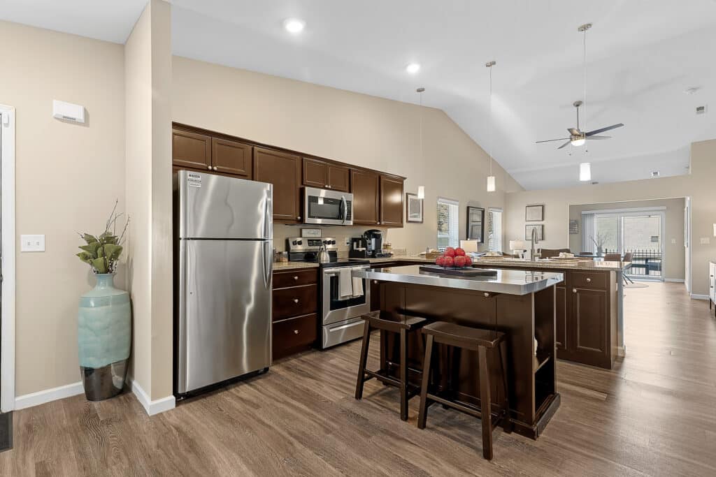 Modern kitchen with stainless steel appliances, dark wood cabinets, island with stools, and a vase with branches next to the entryway; open layout to dining and living areas.