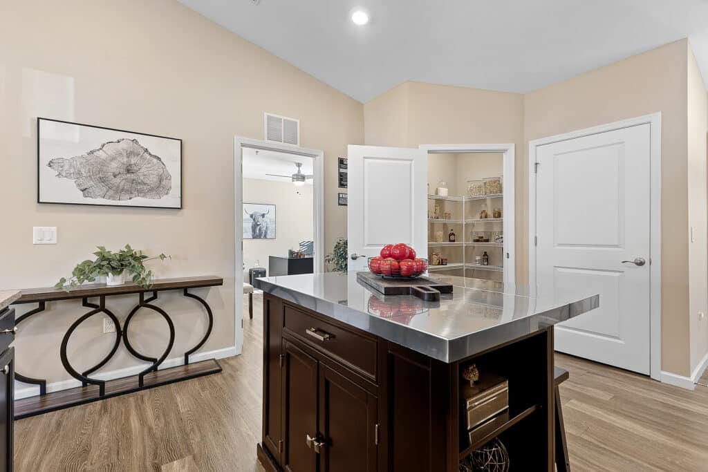 Modern kitchen with a stainless steel island, wooden floors, open pantry door, closed white door, console table, and wall art. A bowl of red apples is on the island.