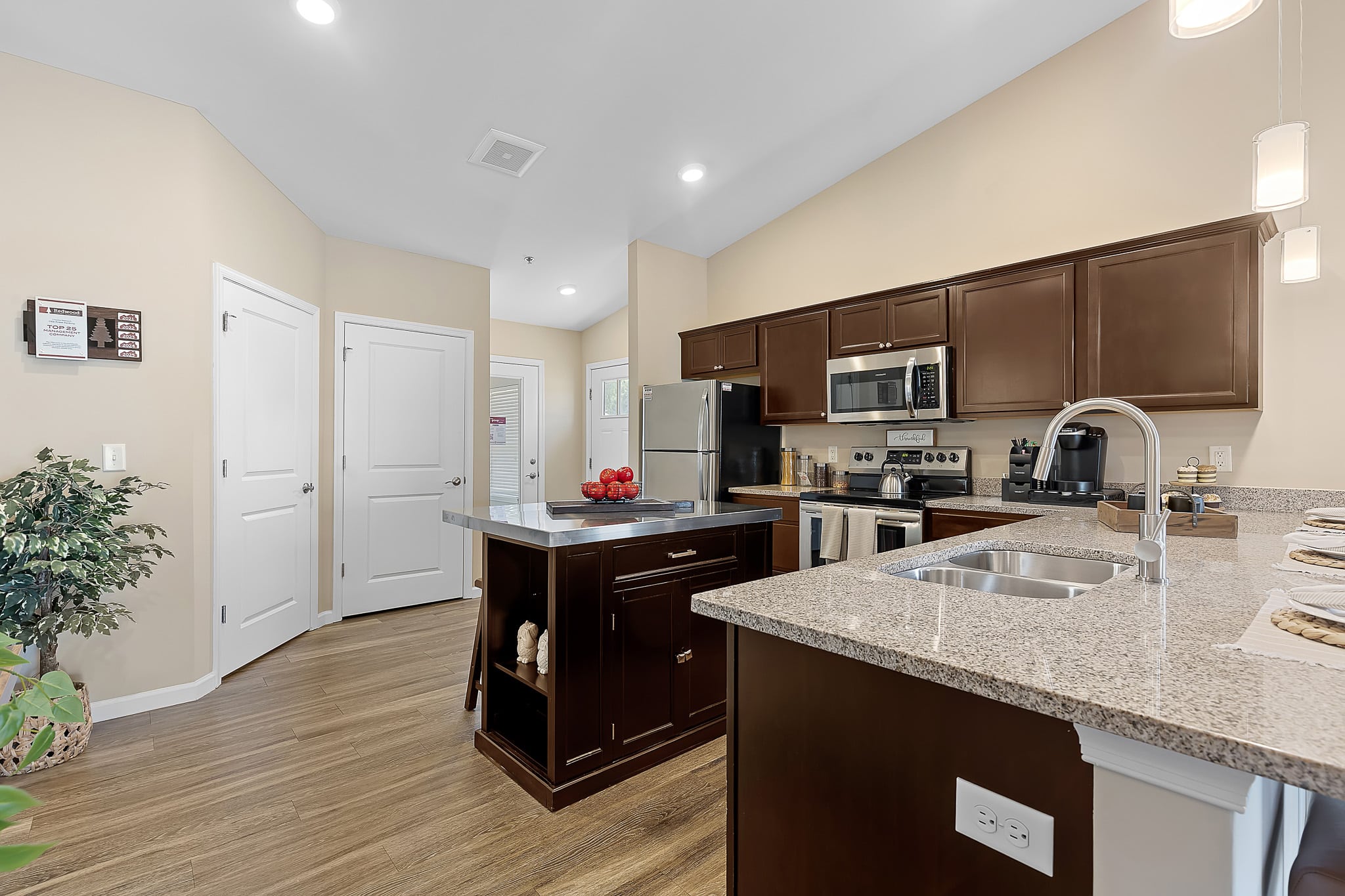 Modern kitchen with dark wood cabinets, stainless steel appliances, granite countertops, center island, and wood flooring. Three pendant lights hang above the counter.