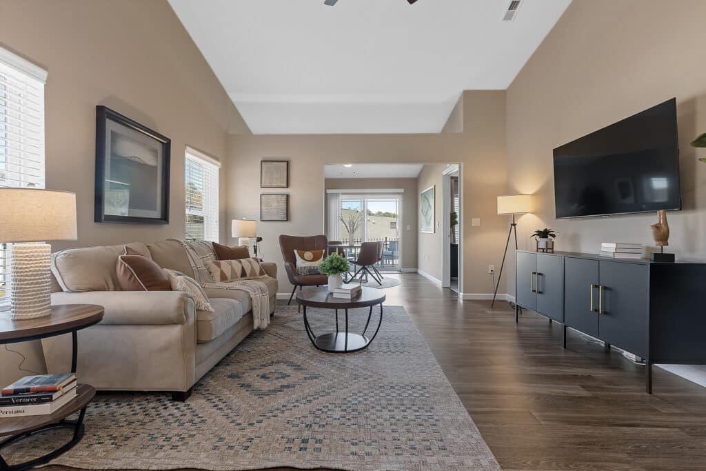 A modern living room with a beige sofa, round coffee table, large rug, wall-mounted TV, and sideboard, featuring wood flooring and large windows letting in natural light.