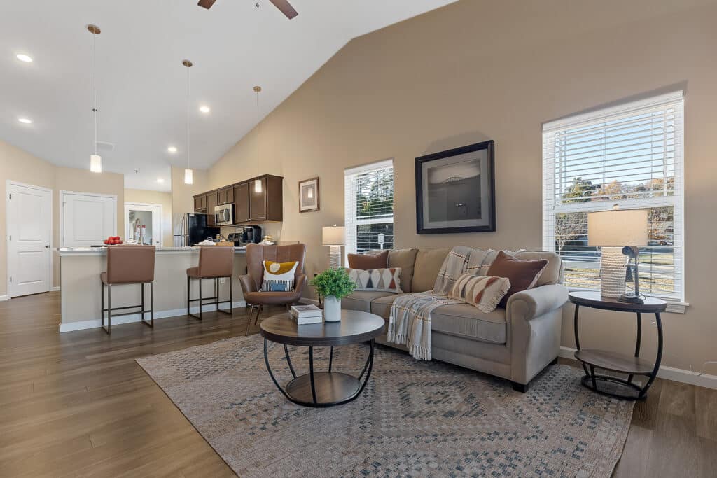 A modern open-concept living room and kitchen with neutral decor, tan sofa, round coffee table, bar stools, pendant lights, and large windows letting in natural light.