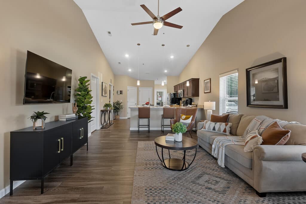 Open-concept living room and kitchen with beige walls, a ceiling fan, wall-mounted TV, sofa, coffee table, and dining area, decorated with plants and framed art.