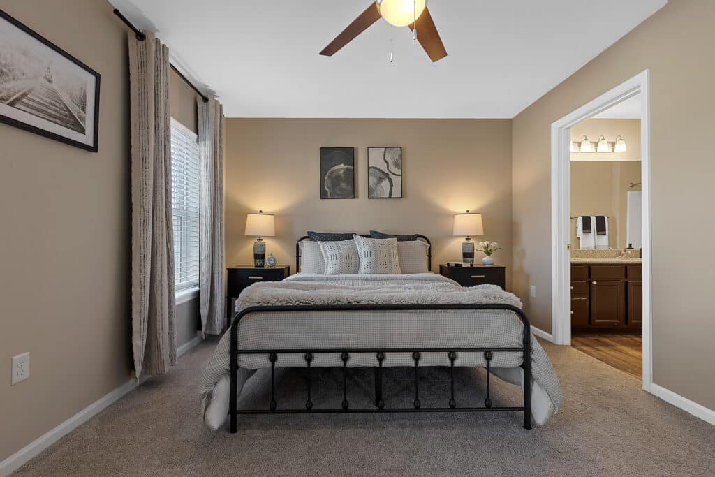 A modern bedroom with a metal bed frame, neutral bedding, two nightstands with lamps, carpeted floor, and an adjoining bathroom visible through an open doorway.