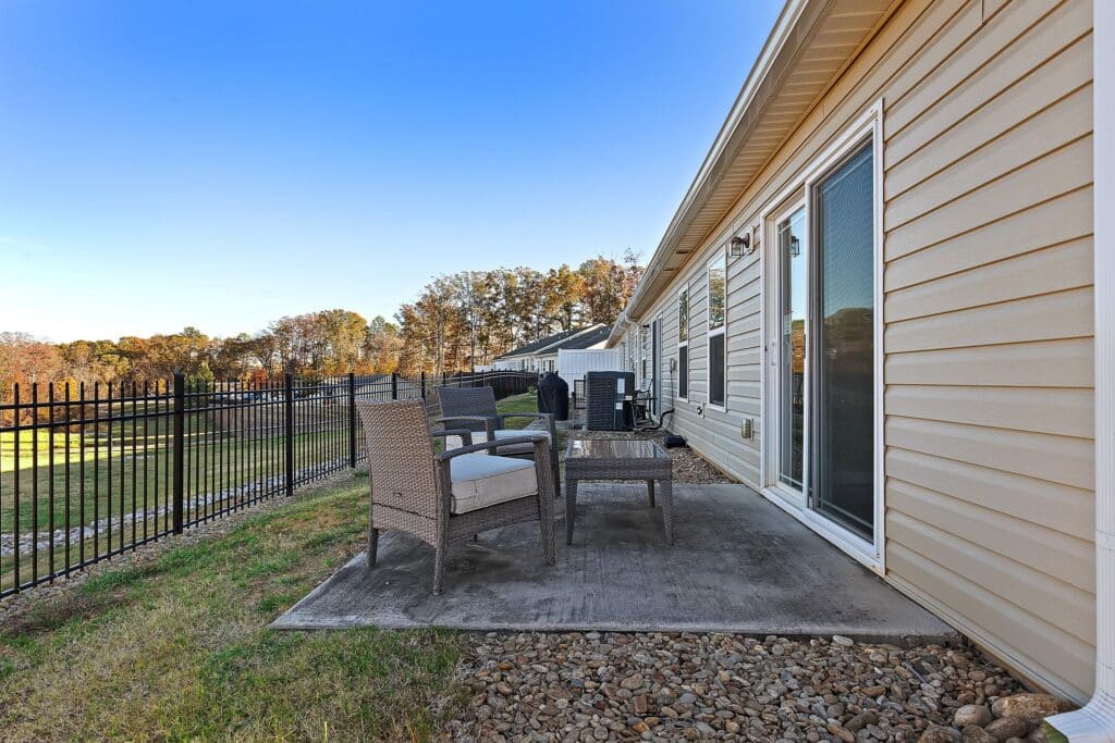 A small concrete patio with wicker chairs and a table sits next to a beige house with sliding glass doors, overlooking a fenced grassy yard.