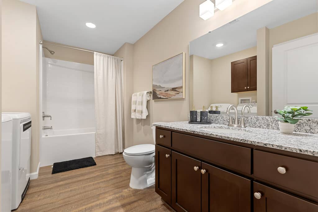 Modern bathroom with granite countertop, dark wood vanity, toilet, bathtub with shower curtain, light wood flooring, and neutral walls decorated with towel and framed art.