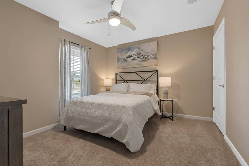 A bedroom with beige walls, a bed with white bedding, two bedside tables with lamps, a ceiling fan, a window with curtains, and a landscape painting above the bed.