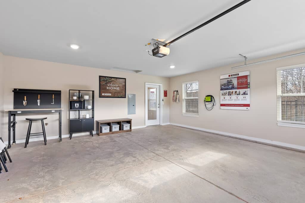 A clean, organized garage with beige walls, storage shelves, a black tool cabinet, posters on the walls, and natural light coming through the windows.