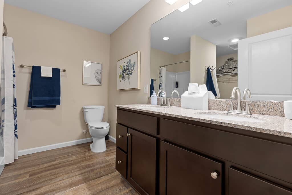 A modern bathroom with double sinks, dark wood cabinets, a toilet, towel rack with blue towels, wall art, and a shower with a white curtain.