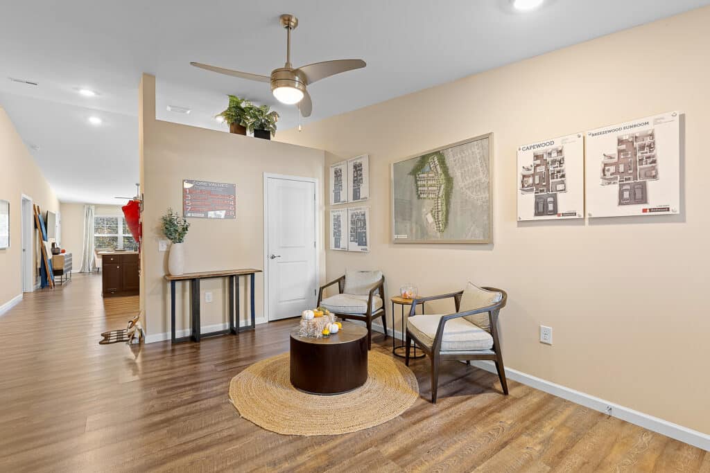 A small seating area with two chairs and a round table on a rug, wall art and maps, wood floor, and a hallway leading to other rooms in a modern interior.