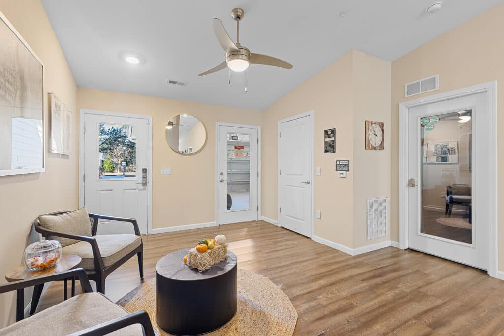 A waiting room with two chairs, a round table with autumn decor, wood flooring, beige walls, a ceiling fan, and several doors including one with a glass panel.