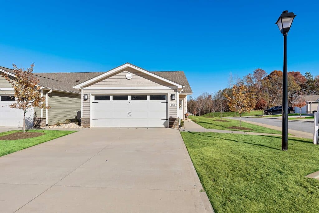 Single-story house with a two-car garage, concrete driveway, and well-maintained lawn on a suburban street with trees and a street lamp.