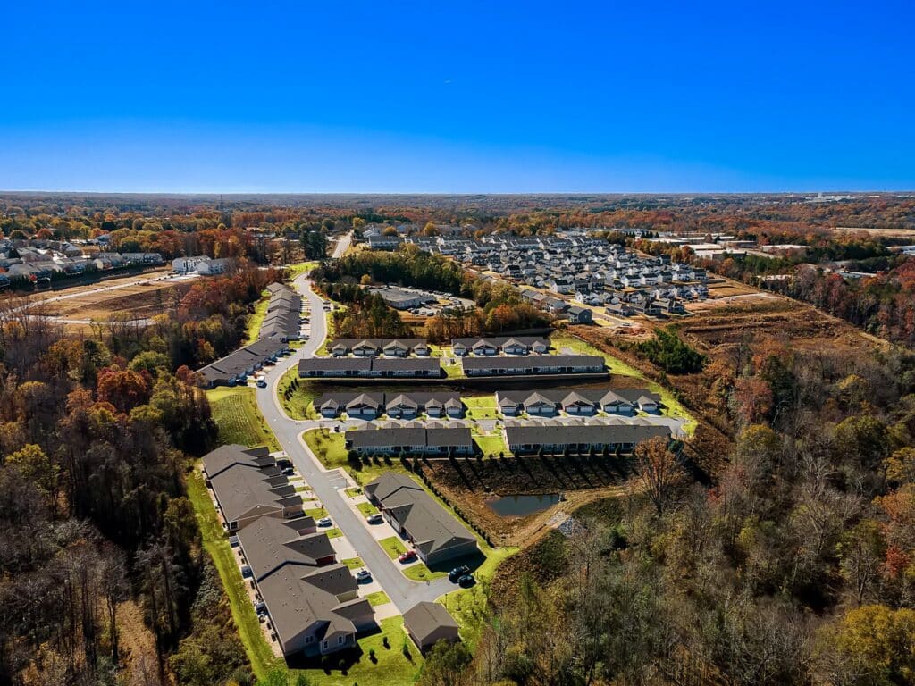 Aerial view of a suburban neighborhood with rows of houses, green lawns, roads, and surrounding trees under a clear blue sky.
