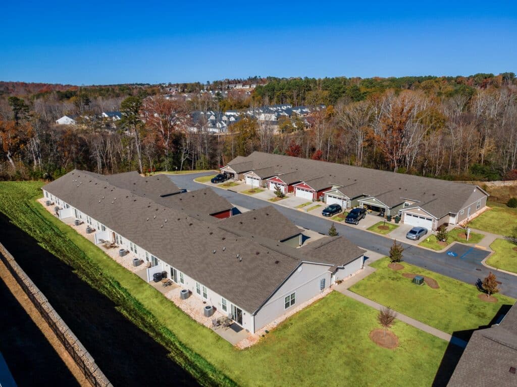 Aerial view of a residential complex with two long, single-story buildings, parking spaces, and landscaped green areas, surrounded by trees and houses in the background.