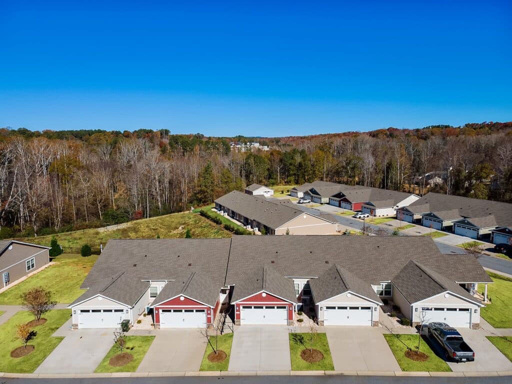 A row of connected single-story townhouses with garages, driveways, and cars parked in front, with trees and additional houses in the background under a clear blue sky.