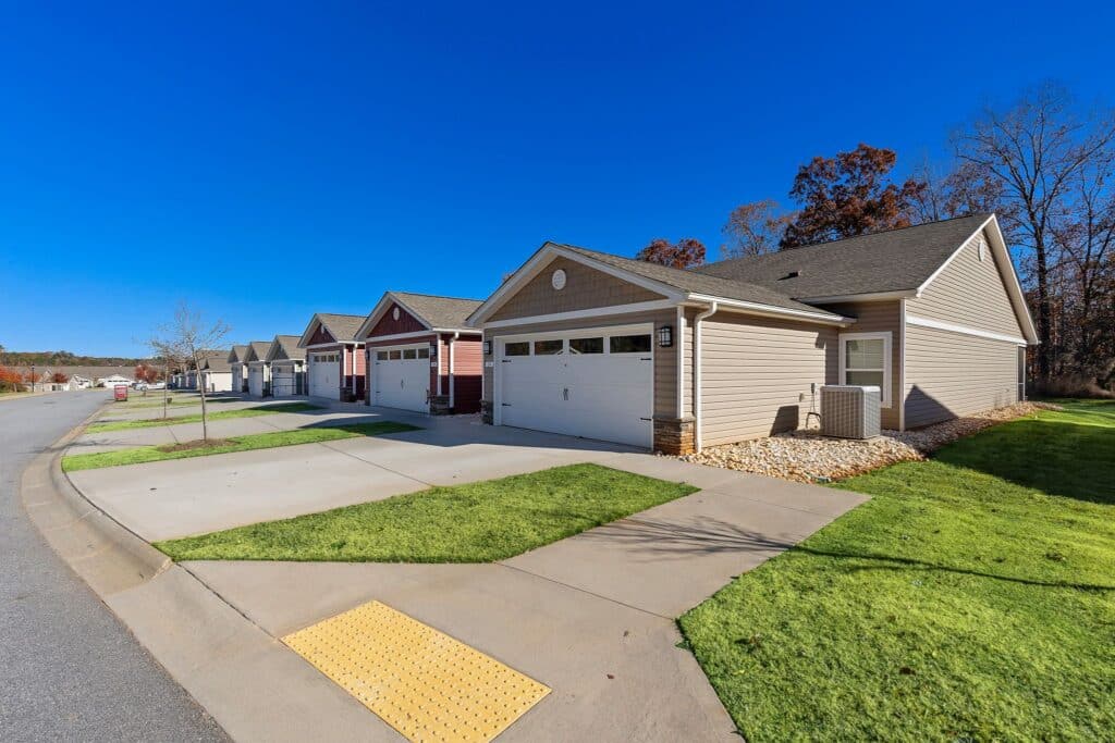 Row of single-story suburban houses with attached garages and driveways, situated along a clean, curved street under a clear blue sky.