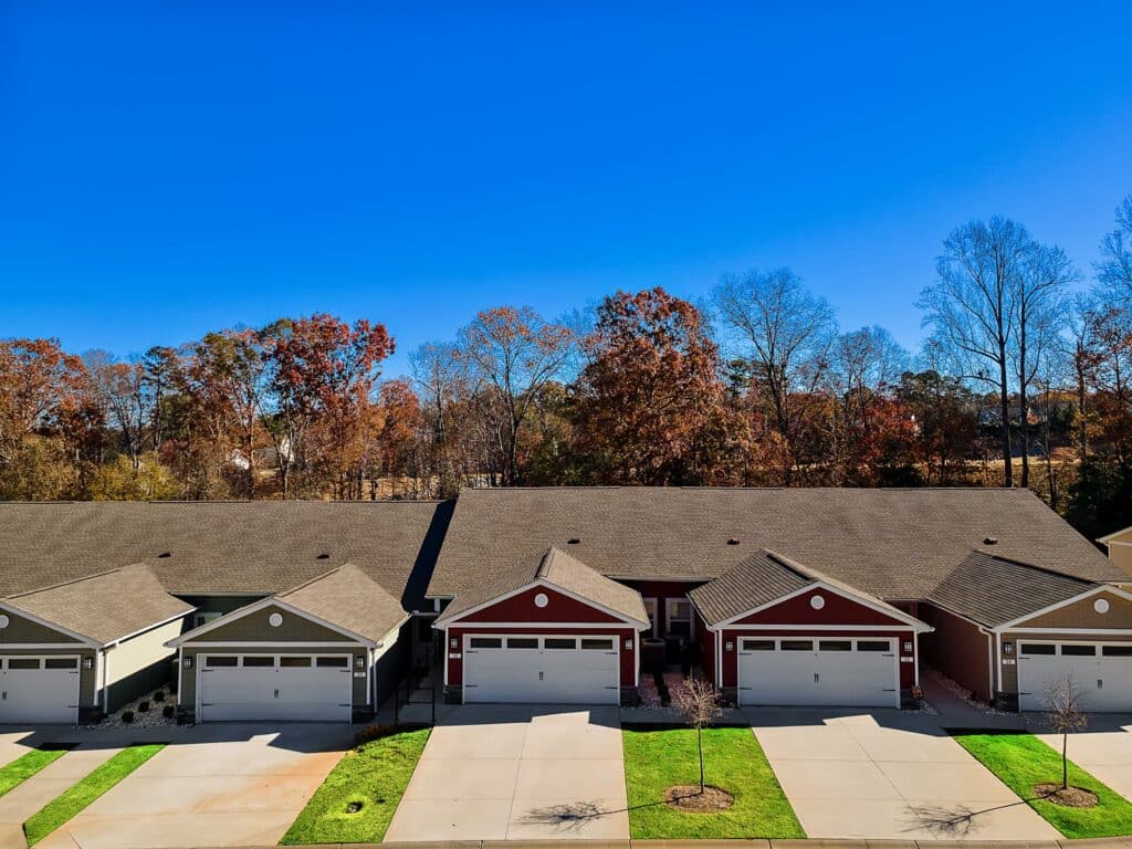 Row of attached single-story townhouses with garages, green lawns, and autumn trees in the background under a clear blue sky.