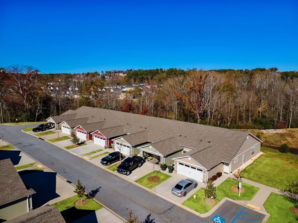 A row of single-story townhouses with driveways and parked cars, bordered by trees under a clear blue sky.