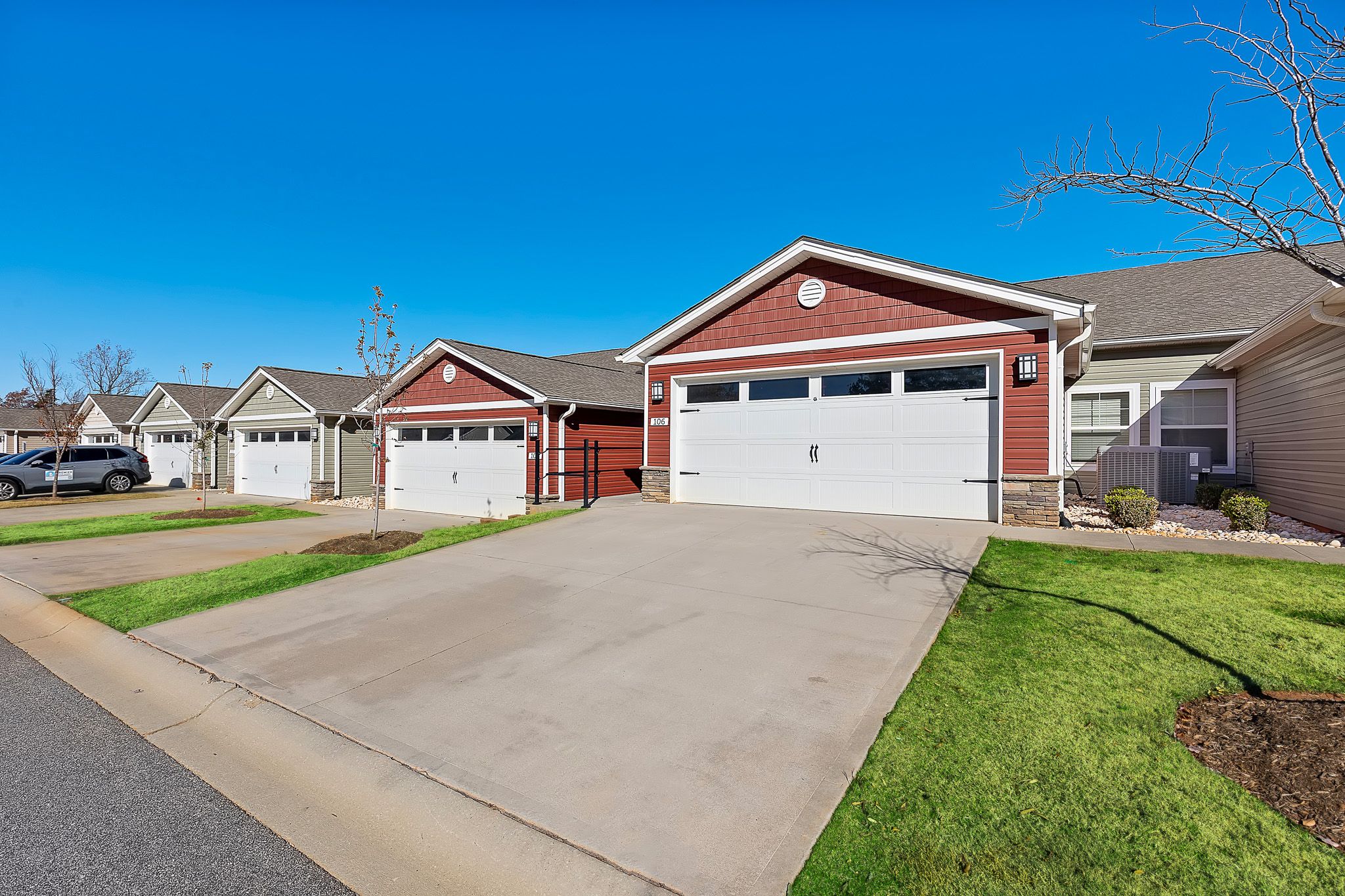 Row of attached single-story homes with red and beige exteriors, each featuring a two-car garage and concrete driveway under a clear blue sky.