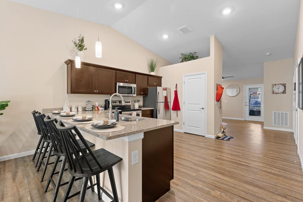 Modern kitchen with dark cabinets, stainless steel appliances, and a breakfast bar with four black chairs; open layout with wood flooring and neutral walls.