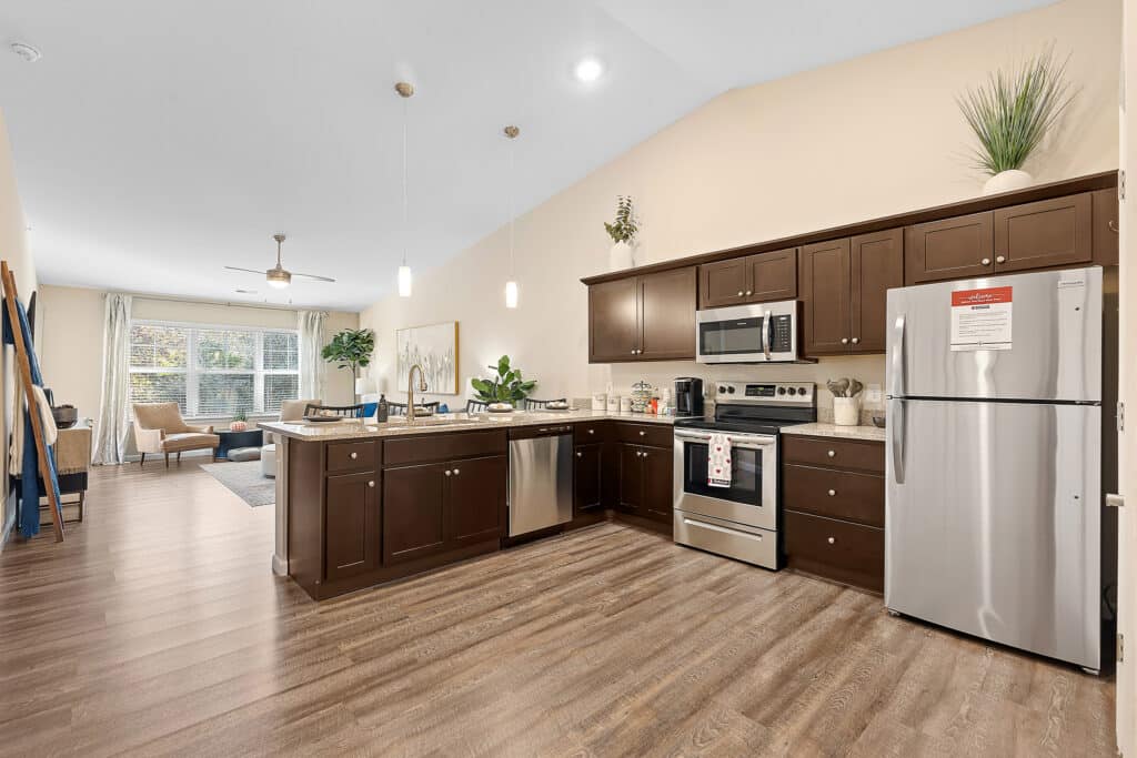 Modern kitchen with dark cabinets, stainless steel appliances, and wood flooring, connected to a bright living area with large windows and neutral decor.