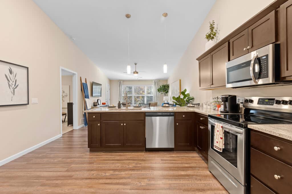 Modern kitchen with dark wood cabinets, stainless steel appliances, and a view into the dining and living area with large windows and hardwood floors.