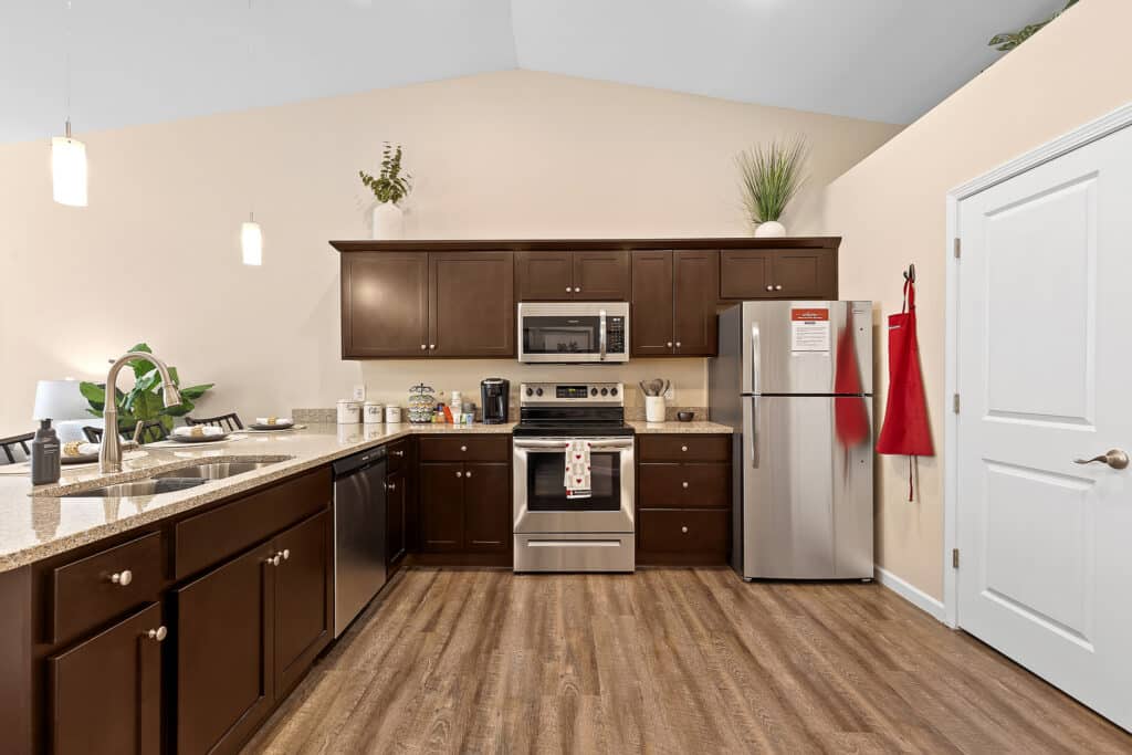 Modern kitchen with dark wood cabinets, stainless steel appliances, granite countertops, and pendant lighting. A red apron hangs on the refrigerator door.