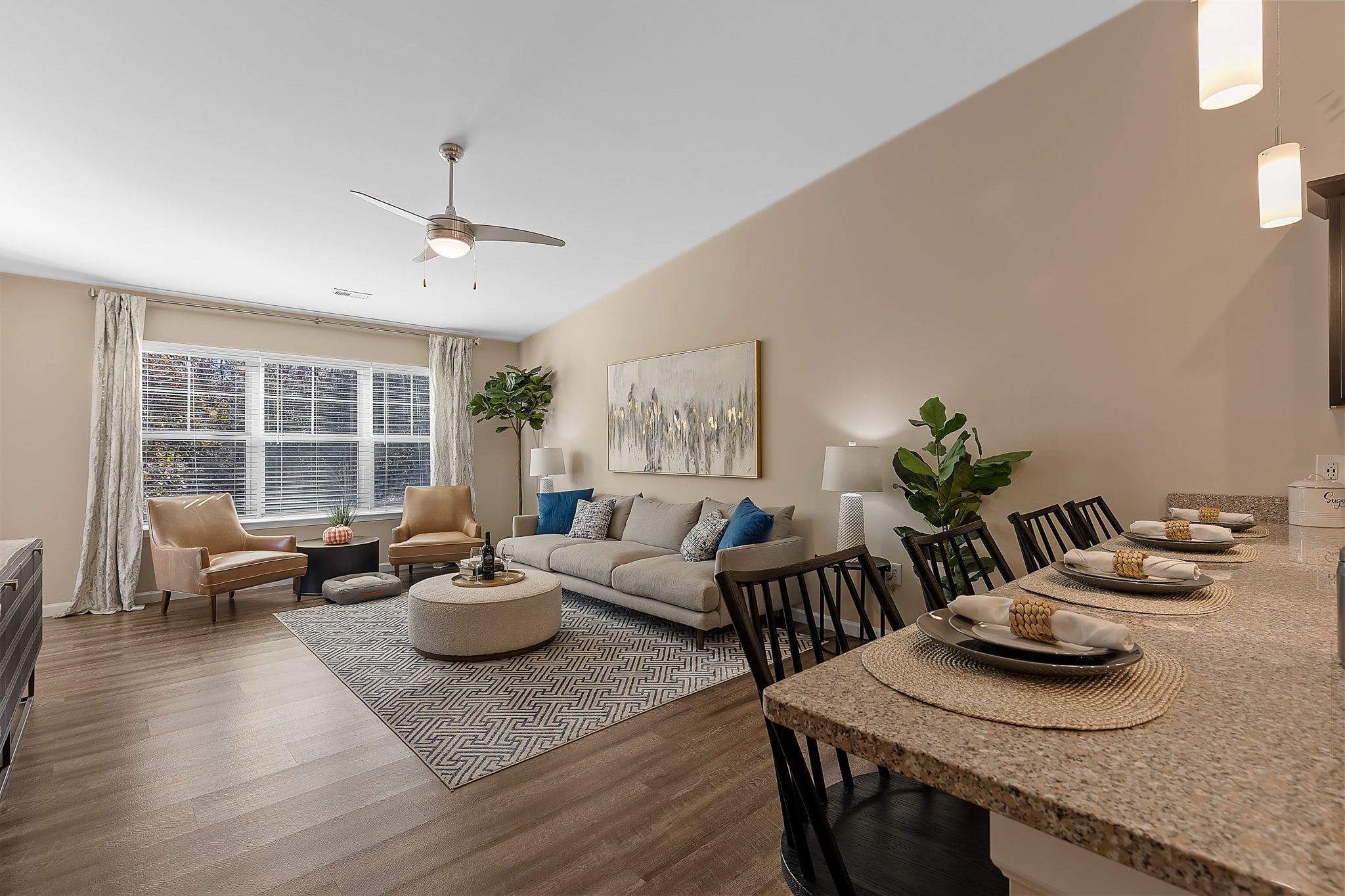 Modern living room and kitchen area with neutral decor, a gray sofa, two beige chairs, a large window, indoor plants, and a granite-topped breakfast bar set with dishes.
