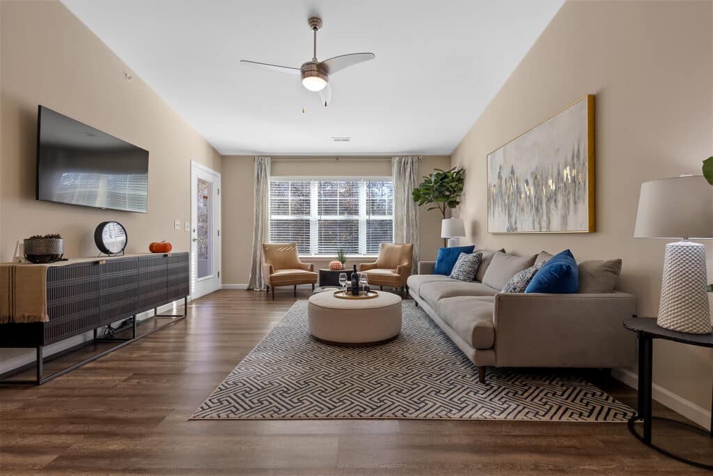 Modern living room with a gray sofa, two beige chairs, a patterned rug, a wall-mounted TV, and large windows letting in natural light.