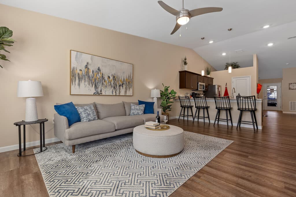Modern living room with beige walls, a sofa, blue pillows, round ottoman, patterned rug, and a kitchen with barstools in the background. Ceiling fan and wall art are visible.