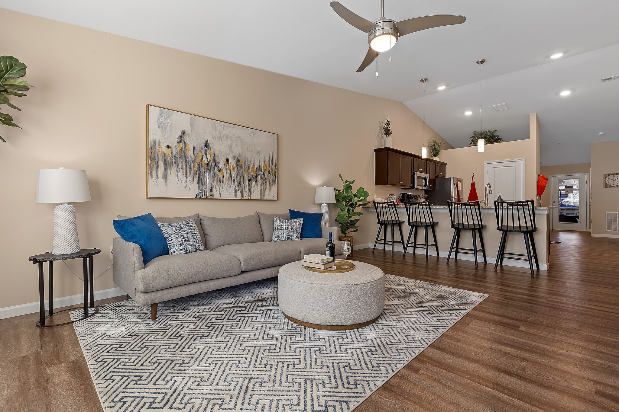 Modern living room with beige walls, a sofa, blue pillows, round ottoman, patterned rug, and a kitchen with barstools in the background. Ceiling fan and wall art are visible.