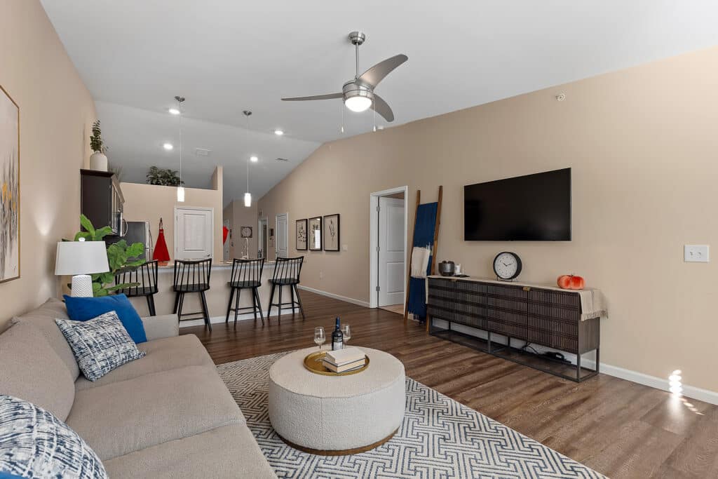 Modern open-concept living room with beige walls, sectional sofa, round ottoman, wall-mounted TV, kitchen bar seating, and hardwood floors.