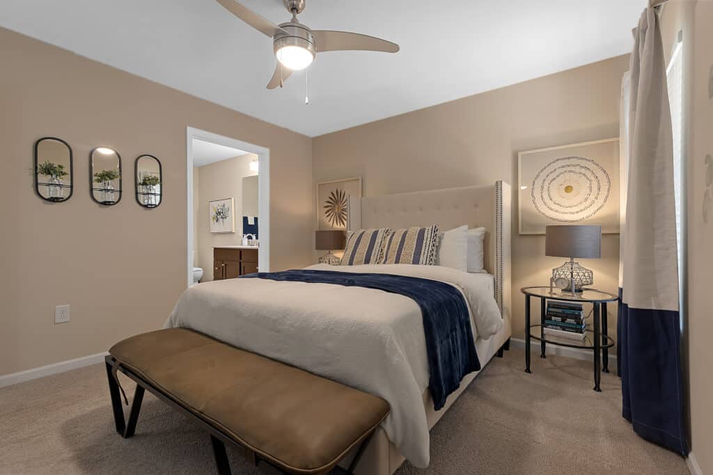 A neatly made bed with blue and white bedding sits in a beige bedroom with a ceiling fan, wall art, a small nightstand, and a bench at the foot of the bed. A bathroom is visible in the background.